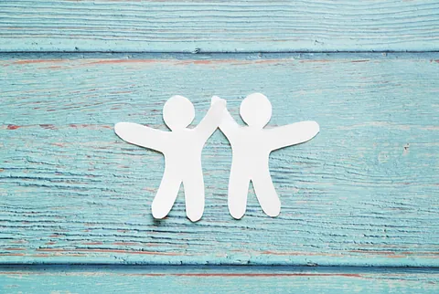 Weißer Papierschnitt von zwei Figuren, die auf blauen Holzbrettern Hände halten.
