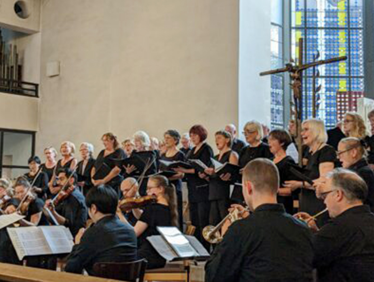 Ein Kirchenchor und Orchester bei einem Auftritt, mit einem Kreuz im Hintergrund.